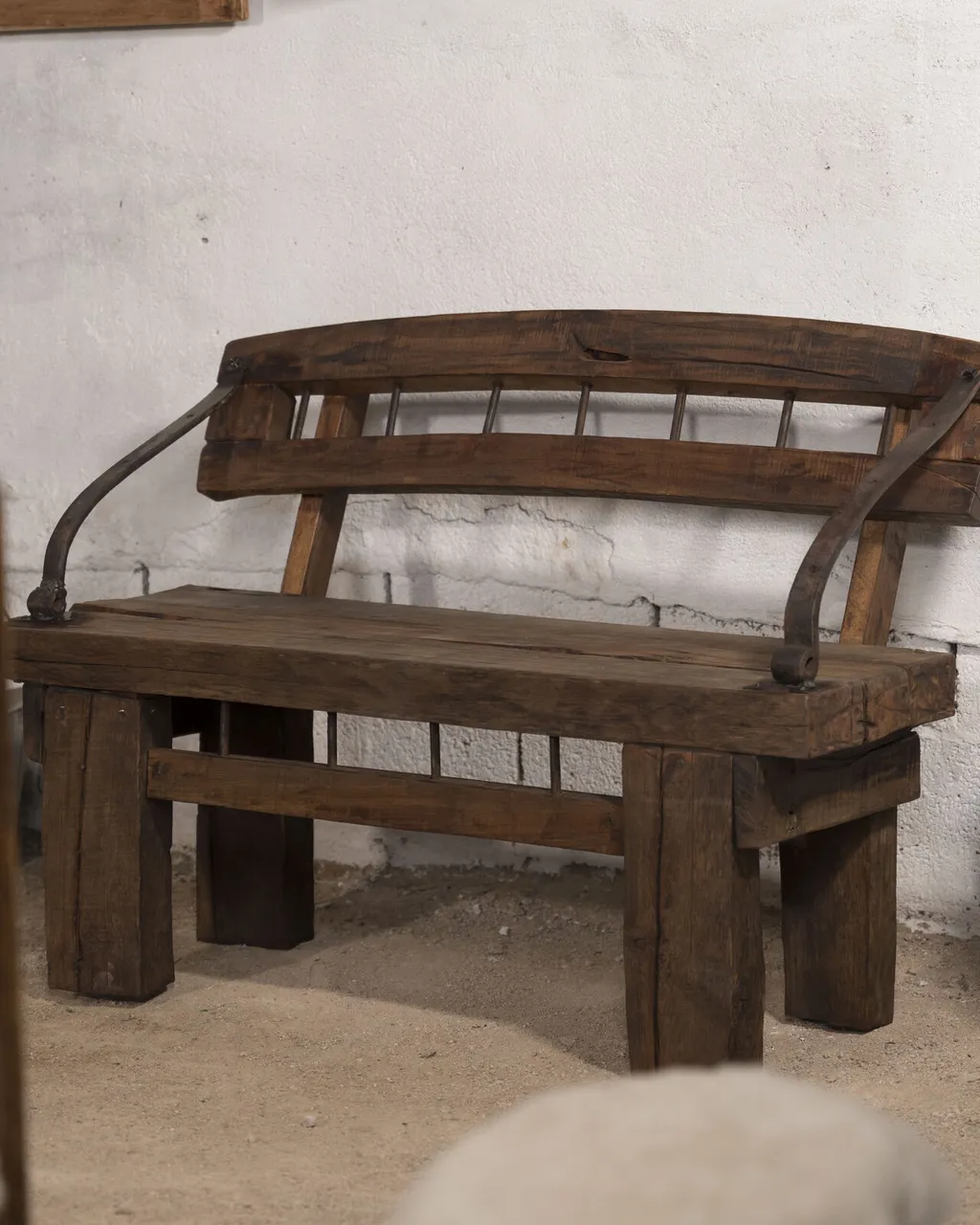 Banco de madera oscura con respaldo de barrotes y brazos de hierro curvado, junto a una lechera